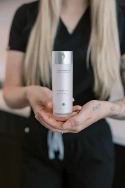 A person in black scrubs holding a bottle of AnteAGE MD Accelerator skin moisturizer.